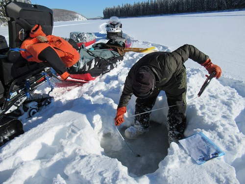A scientist dressed in a puffy jacket, bibs, bunny boots, and rubber gloves leans over a small hole drilled in river ice. He is holding a measuring tape that extends into the hole. There are two snowmachines with sleds of gear visible in the background.