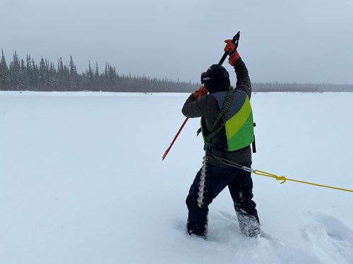 A man stands on a snow-and-ice covered river in a life jacket holding an ice spud above his head, preparing to strike the ice to test the thickness.