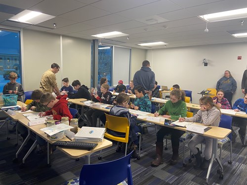 Middle school students sit in three rows of desks in a university classroom, hard at work on their silt stories. Several scientists are chatting with different groups of students.