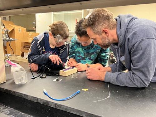 Two students wearing safety goggles and one male scientist are hunched over a small piece of a sensor. One student is using a soldering iron to build the sensor while the other student stabilizes the pieces.