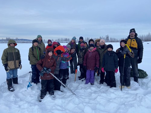 A group of 18 students and several adults stands on a frozen lake covered in snow, dressed in full winter gear. They are standing around an orange stake, which marks the location of a sediment sensor they just installed in the Tanana Lake lagoon.