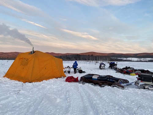 Campsite in the snow. A yellow Arctic Oven tent is in the foreground with smoke coming out of the chimney. Large sleds full of gear are scattered around the campsite. The sky is blue with streaks of clouds.