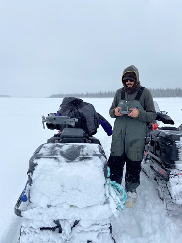 A man in sunglasses and bibs stands next to a snowmachine holding a console for a drone. The drone hovers in the air just about 12 inches over the snowmachine's storage box storage box, ready for takeoff.