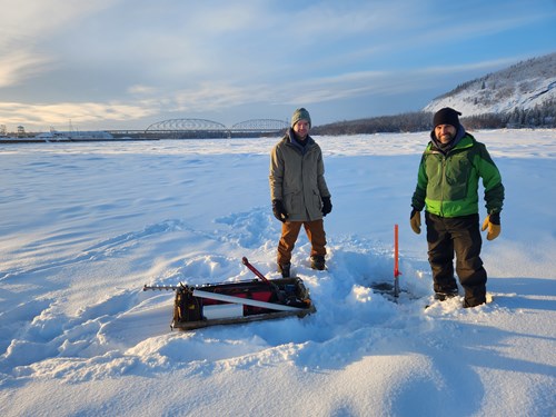Two white men stand on a snow covered, frozen river in full winter gear next to an orange stake that is sticking out of the river ice. The stake marks the location of a sediment sensor in the Tanana River. The Parks Highway bridge over the Tanana River is visible in the background.