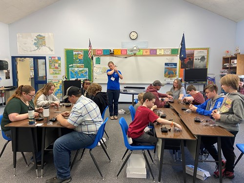Ten students sit in two table groups in a classroom, building toy drones. They receive instructions from a blonde woman scientist standing at the front of the classroom, holding up a drone for demonstration.