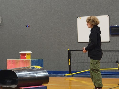 A student wearing a black sweatshirt and army green pants flys a drone through an obstacle course in a school gym. She is attempting to land her drone on a frisbee propped on top of a box jump.