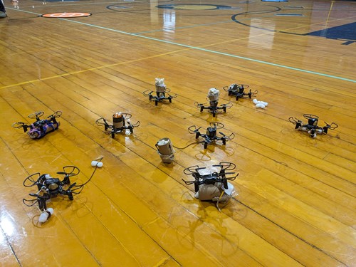 Nine toy drones sit on a school gym floor. Each drone has a different payload system attached, created by the students of Eagle Community School