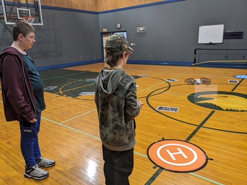 Two students stand with their backs to the camera in a school gym. One student is flying a DJI mini 3 drone while the second student acts as the observer.